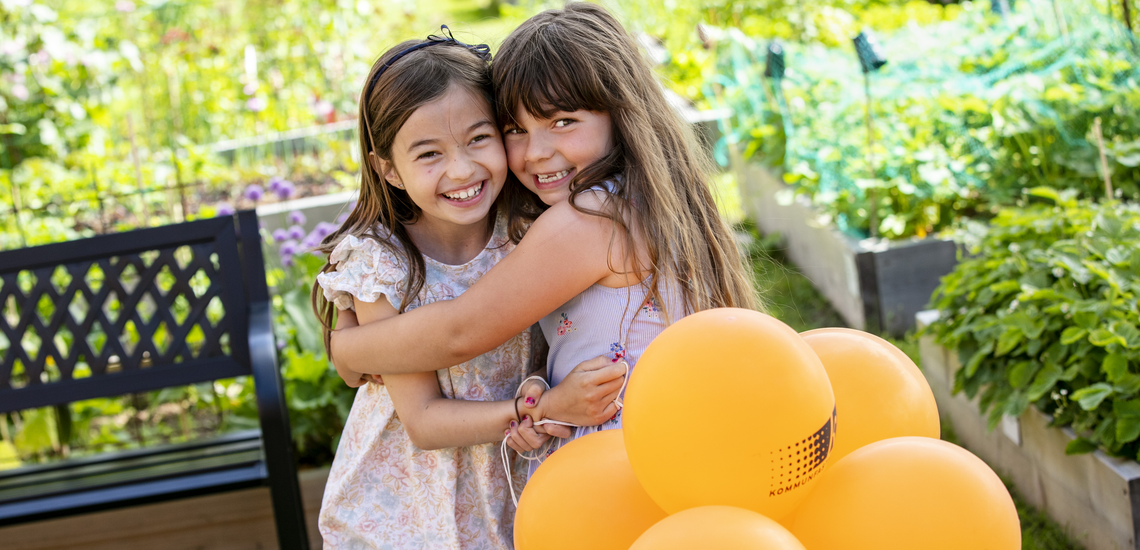 Två tjejer kramar varandra med ballonger en grön sommardag.