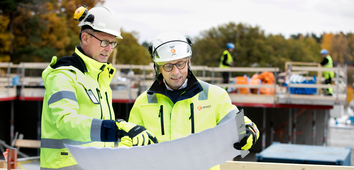 Två byggprojektledare iklädda skyddsutrustning inspekterar en ritning vid byggarbetsplats.