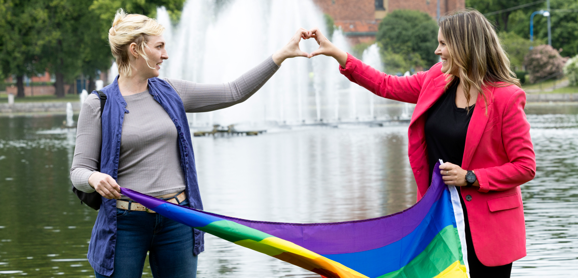 Två kvinnor håller i prideflagga och formar ett hjärta tillsammans med händerna. 