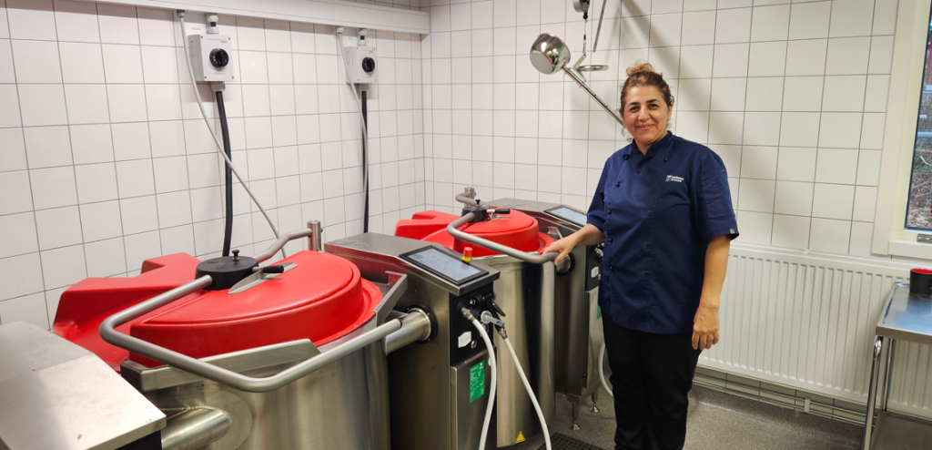 Kvinna står i skolkök intill stora matlagningsgrytor.