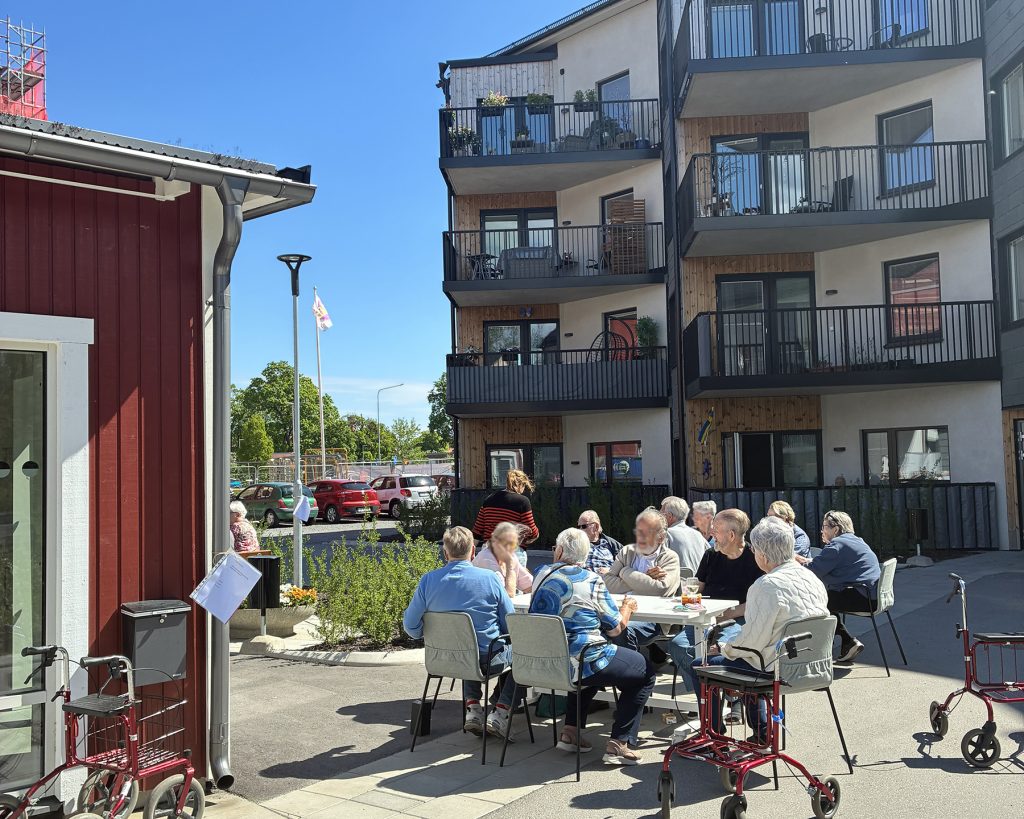 Flera personer sitter på bänkar utanför fastighet och fikar.
