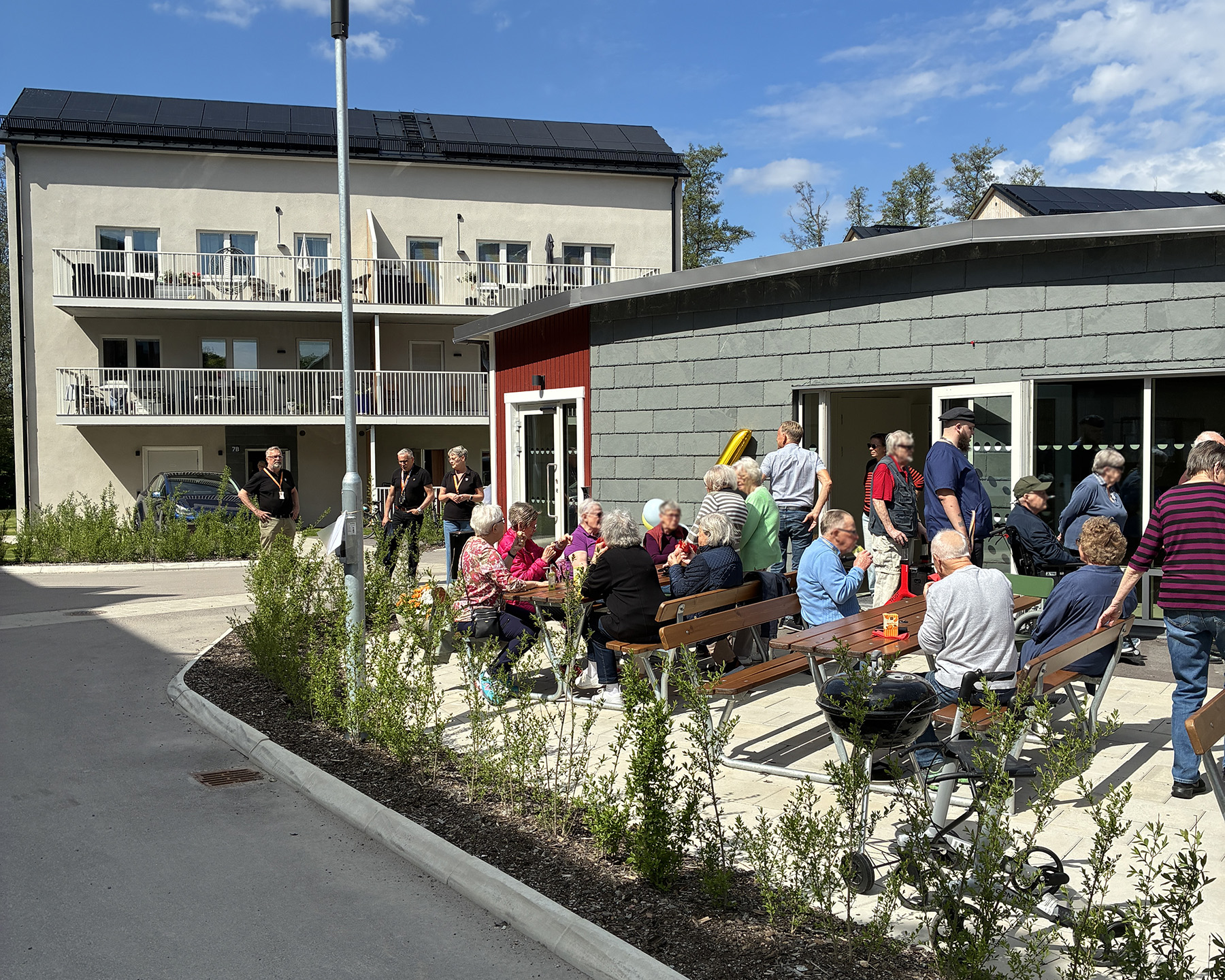 Flera personer sitter på bänkar utanför fastighet och fikar.