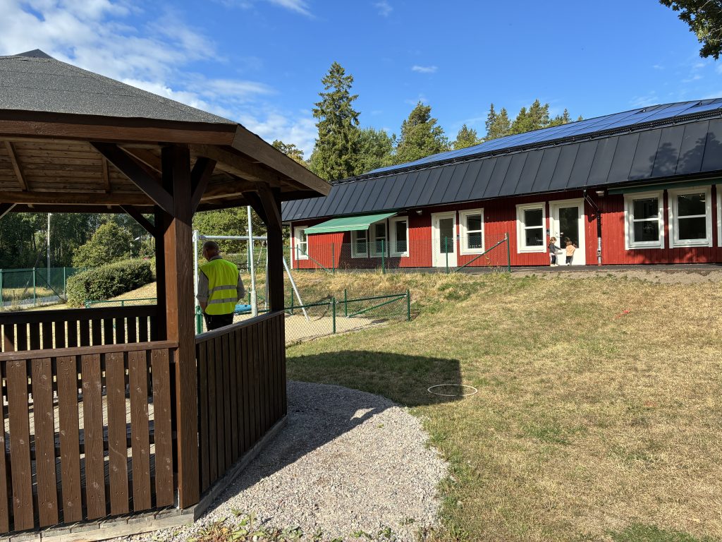 Paviljong med tak utanför röd byggnad.